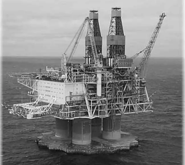 Black and white image of a large offshore oil drilling rig with three supporting columns, cranes, and industrial structures against the sea.