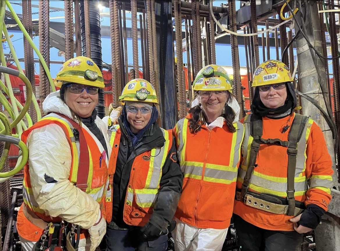 Two construction workers in hard hats are on a scaffolding, managing steel rebar as they work on a building site under a blue sky.